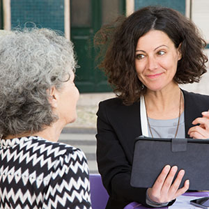 Smiling young consultant offering pen to elderly client for signing documents. Senior grey haired lady talking to young positive woman in jacket with tablet and pen. Agent work concept